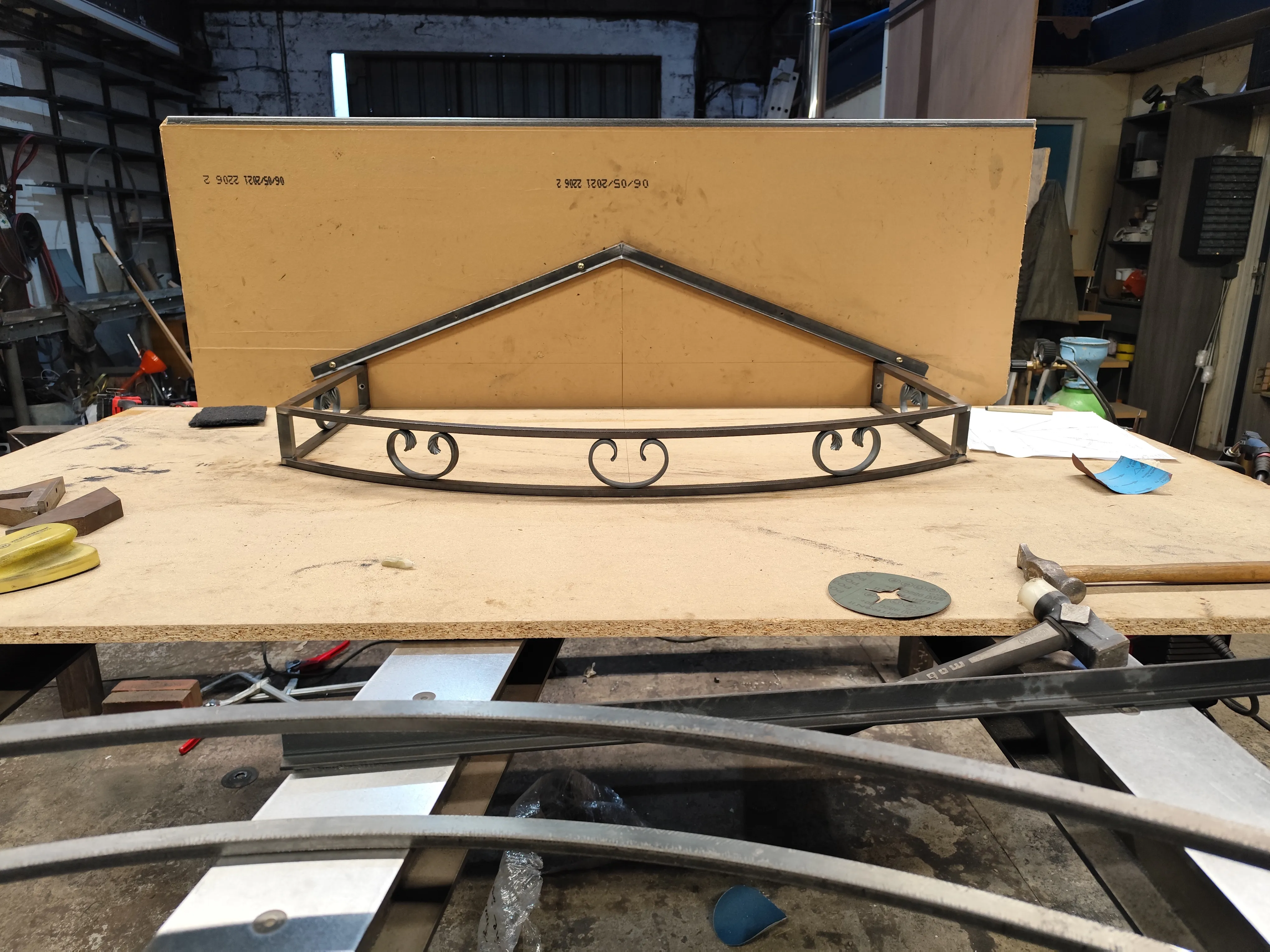 Assemblage de l'armature de la marquise en acier sur établi Armature de toit d'une marquise en acier de forme triangulaire avec une face avant courbée ornée de volutes, posée à plat sur un établi en bois.