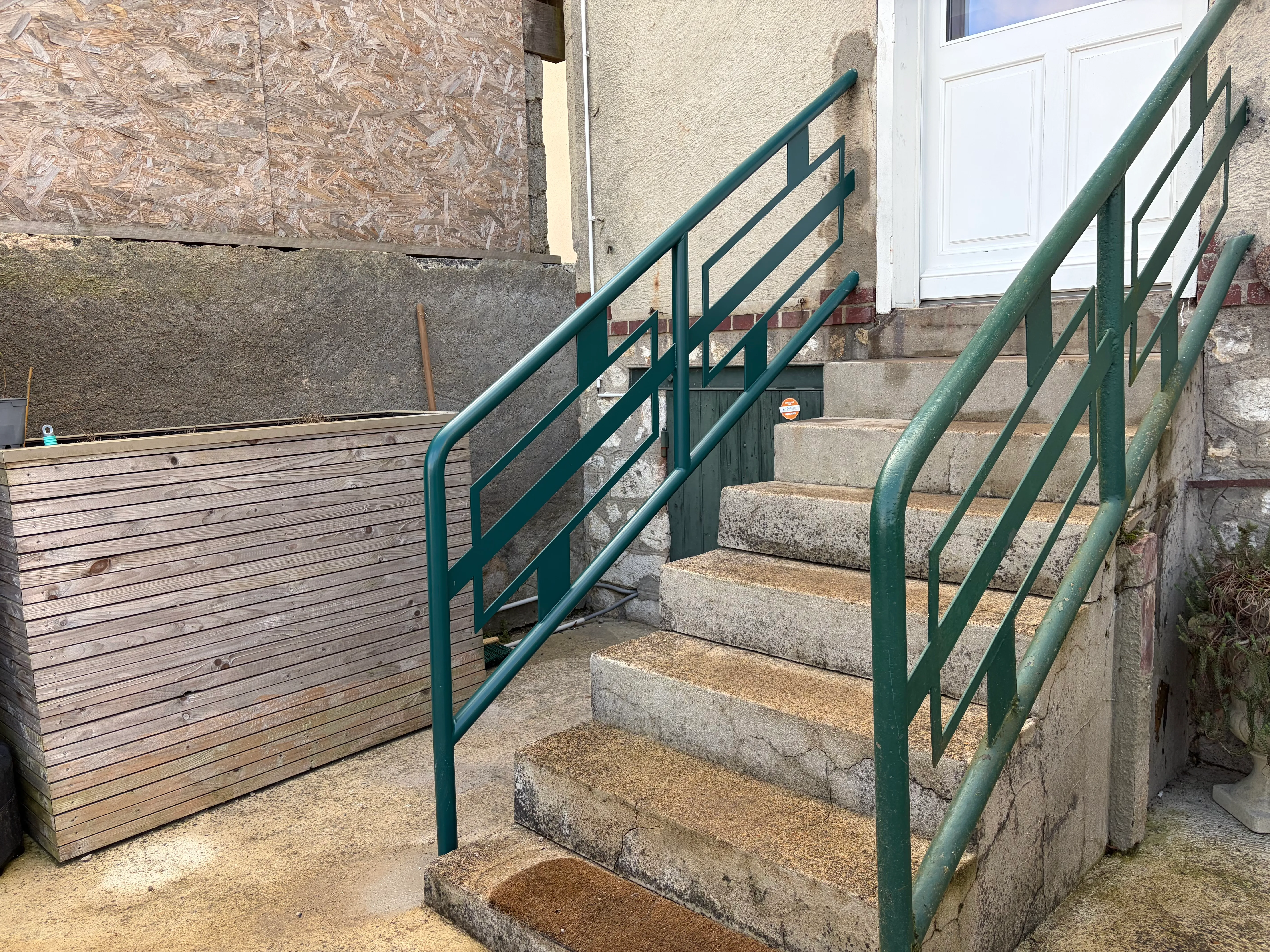 Rampe d'escalier extérieur en acier peint - Ferronnerie à Chartres Rampe d'escalier extérieur en acier avec finition peinture polyuréthane verte anti-rouille. Garde-corps aux motifs géométriques rectangulaires fixé sur un escalier en béton menant à une entrée.