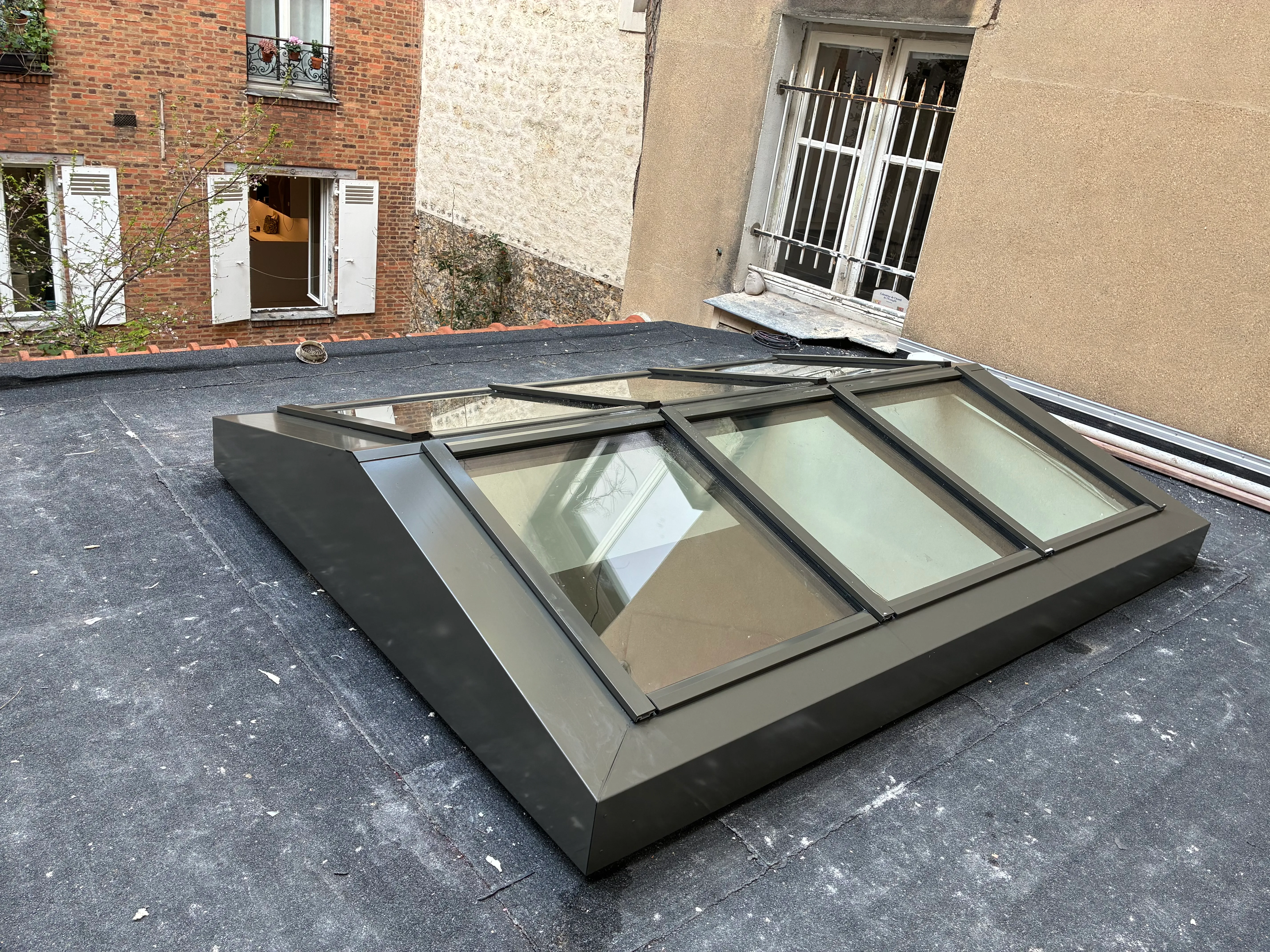 Verrière de toit sur mesure en profilés JANSEN sur toit terrasse à Paris Vue d'ensemble d'une nouvelle verrière de toit à double pente en acier sombre, installée sur un toit terrasse parisien entouré de façades en briques et pierres.