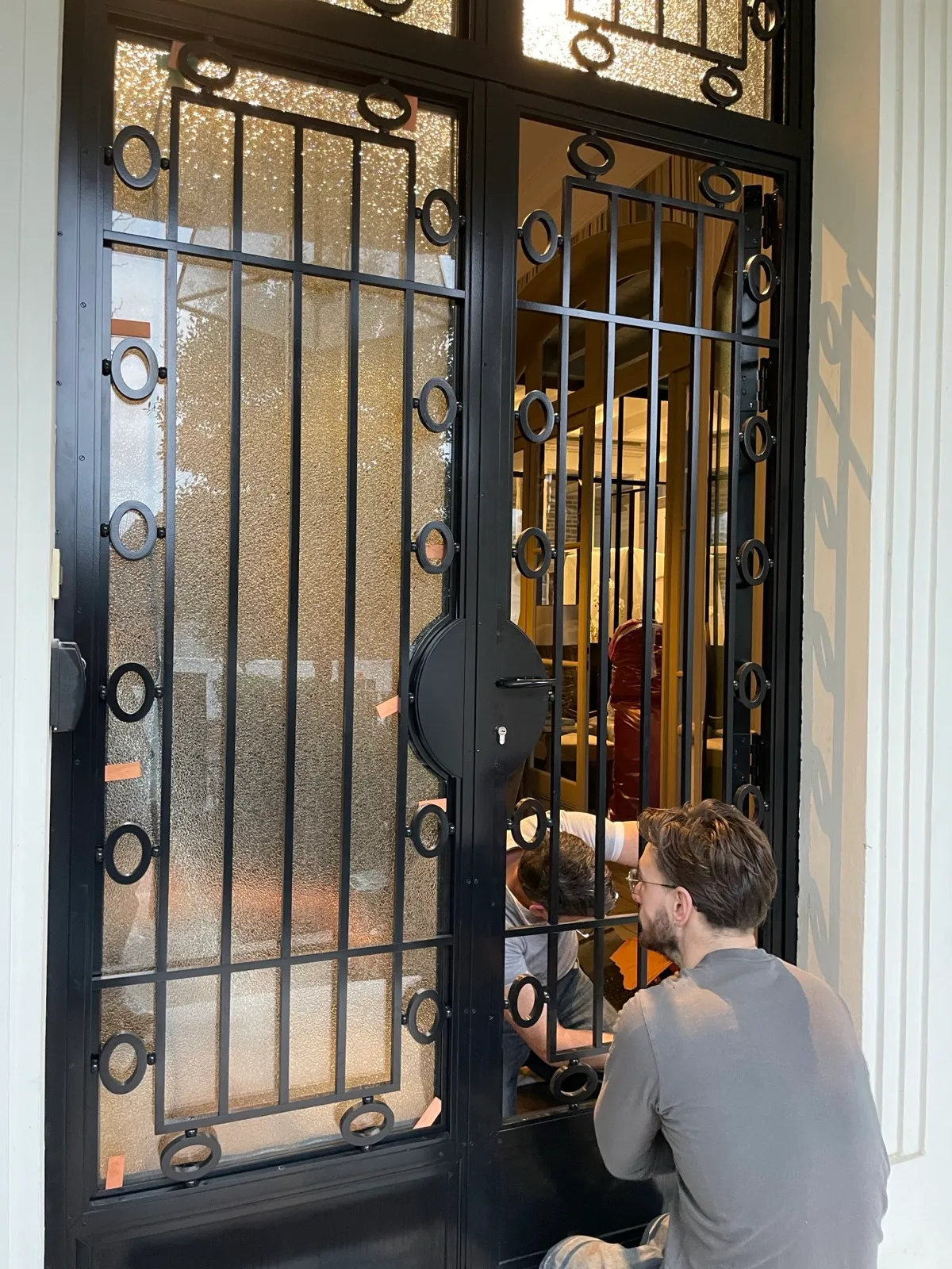 Deux artisans serruriers installant le mécanisme d'une imposante porte à double battant en métal noir ornée de motifs circulaires et de verre texturé