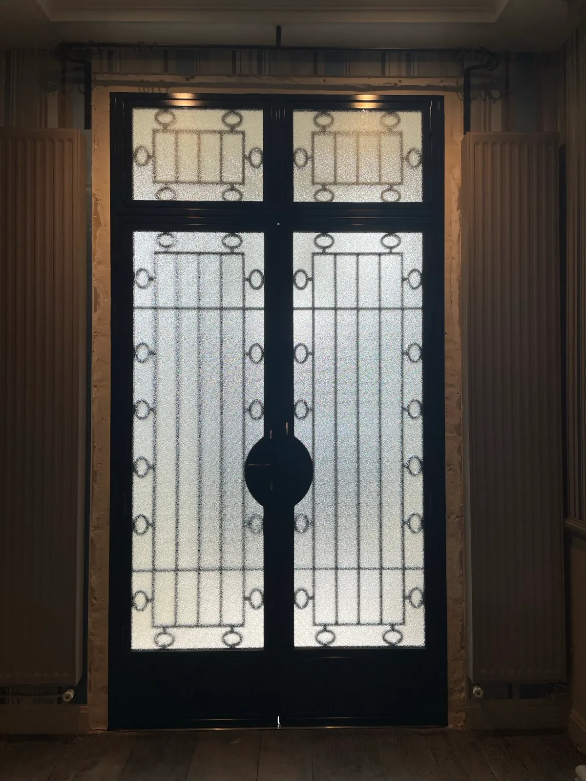 Ouvrages métalliques sur mesure pour architectes : allier esthétique et technique Vue intérieure d'une porte ou cloison métallique vitrée, révélant ses vitrages texturés lumineux, son armature noire structurée et sa grande serrure centrale